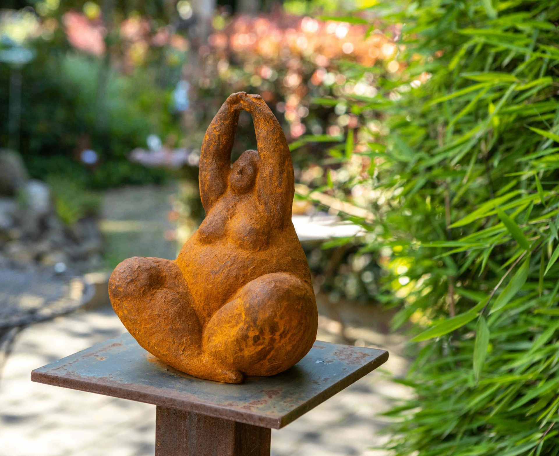 Roostbruine yoga dame tuinbeeld op een metalen sokkel in een groene tuin